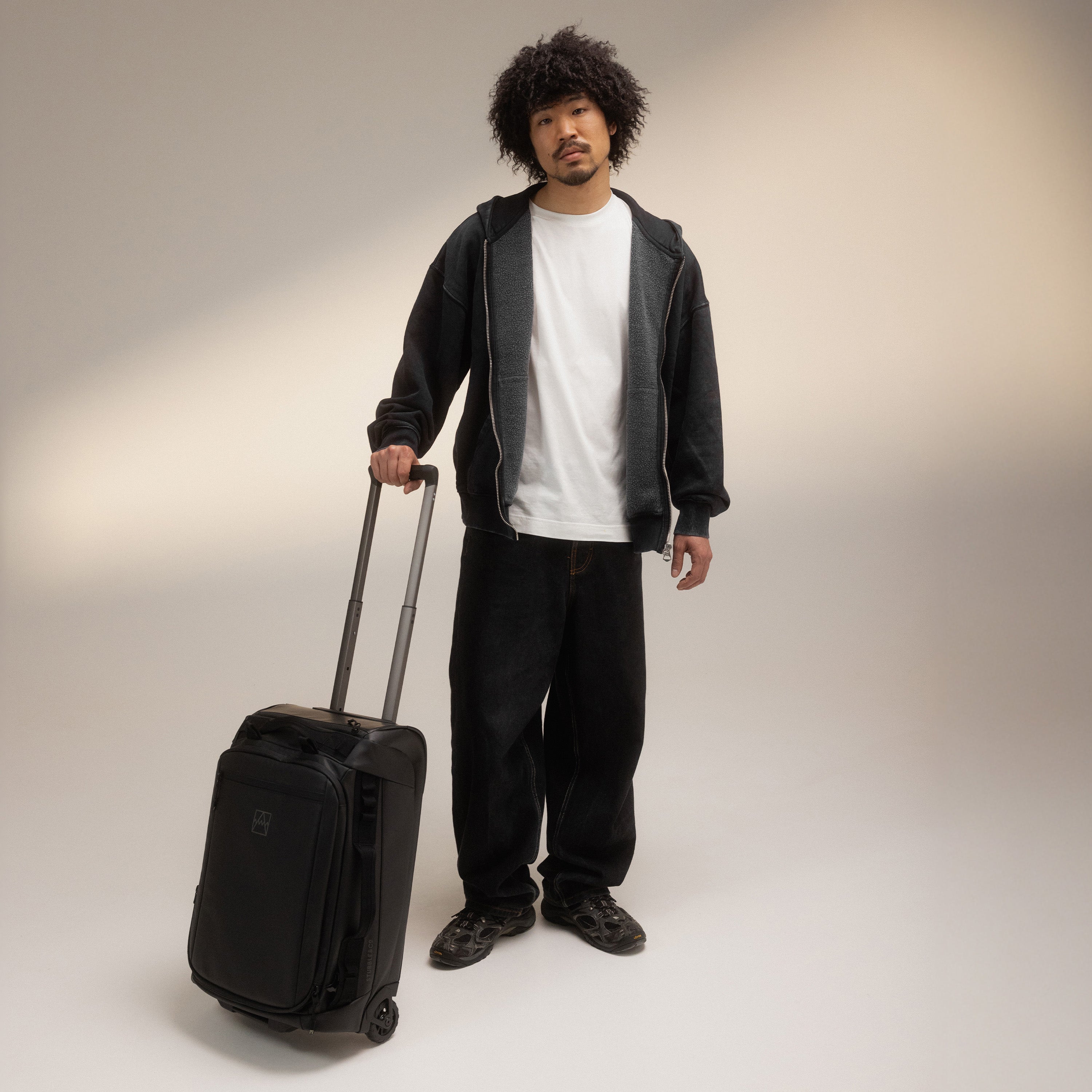 A man standing in a studio holding the handle of the Wheeled Kit Bag 40L in All Black