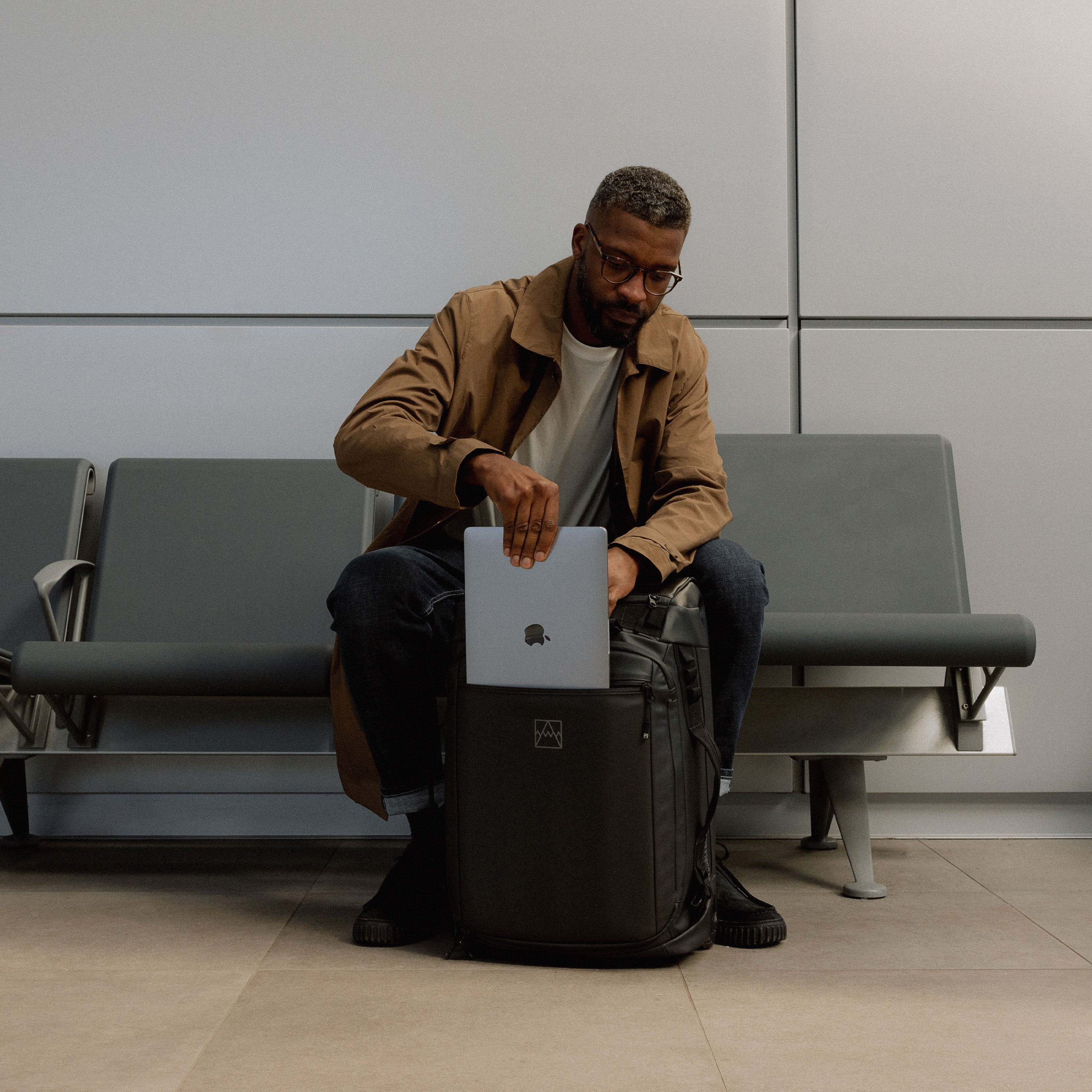 A man sitting on an airport bench putting a laptop into a Wheeled Kit Bag 40L in All Black