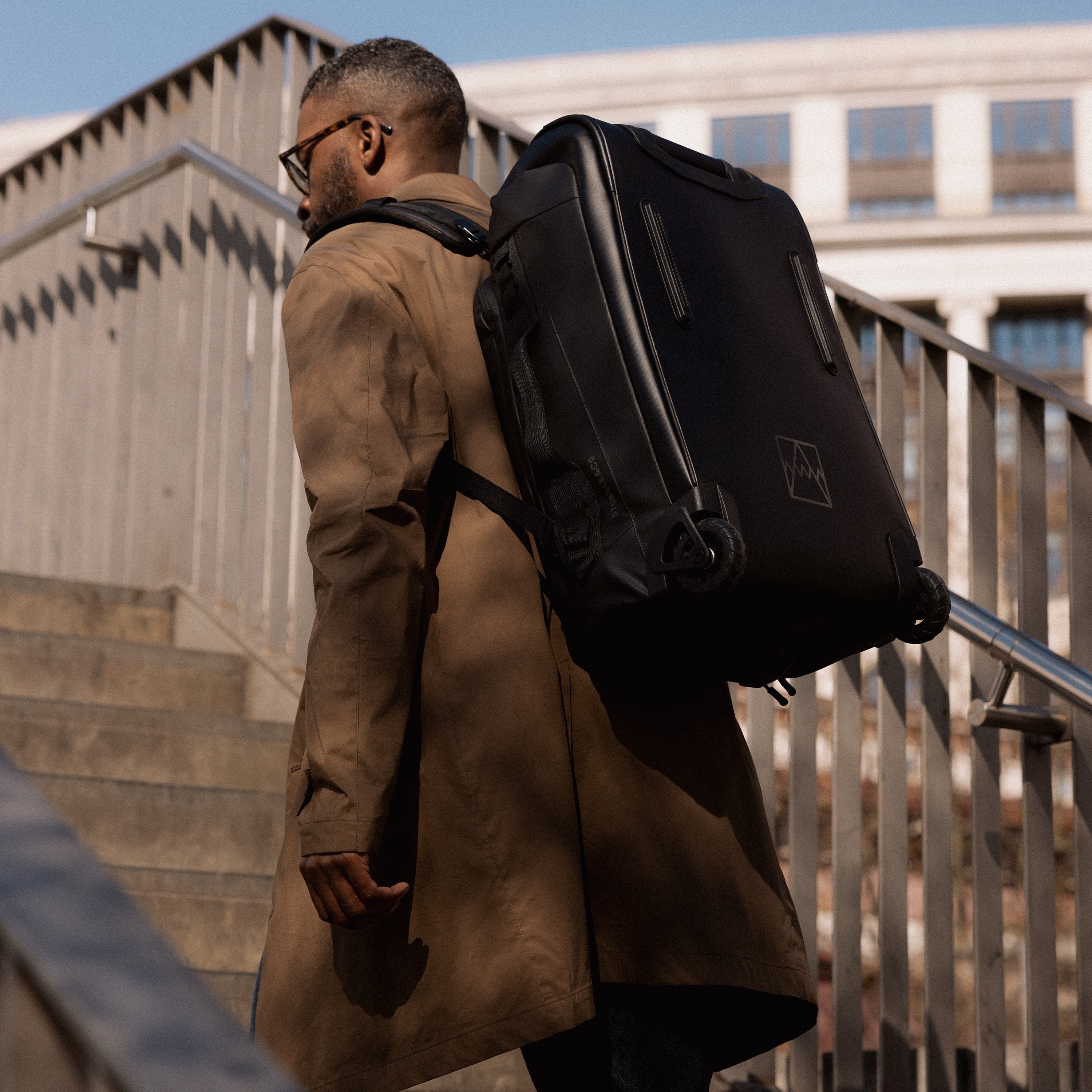 A man walking up the stairs wearing a Wheeled Kit Bag 40L in All Black on his back