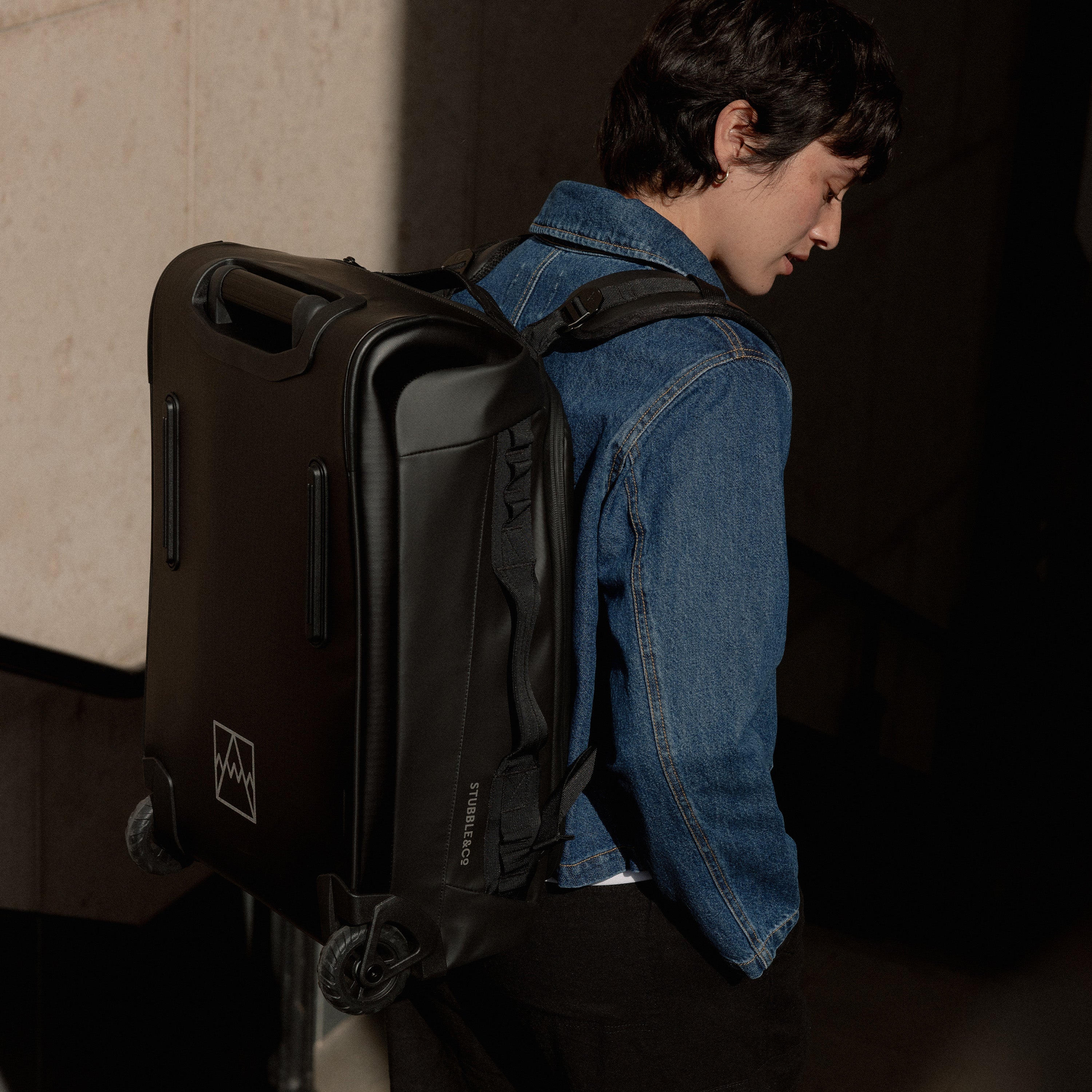 A studio shot of a woman wearing an All Black Wheeled Kit Bag 40L on her back