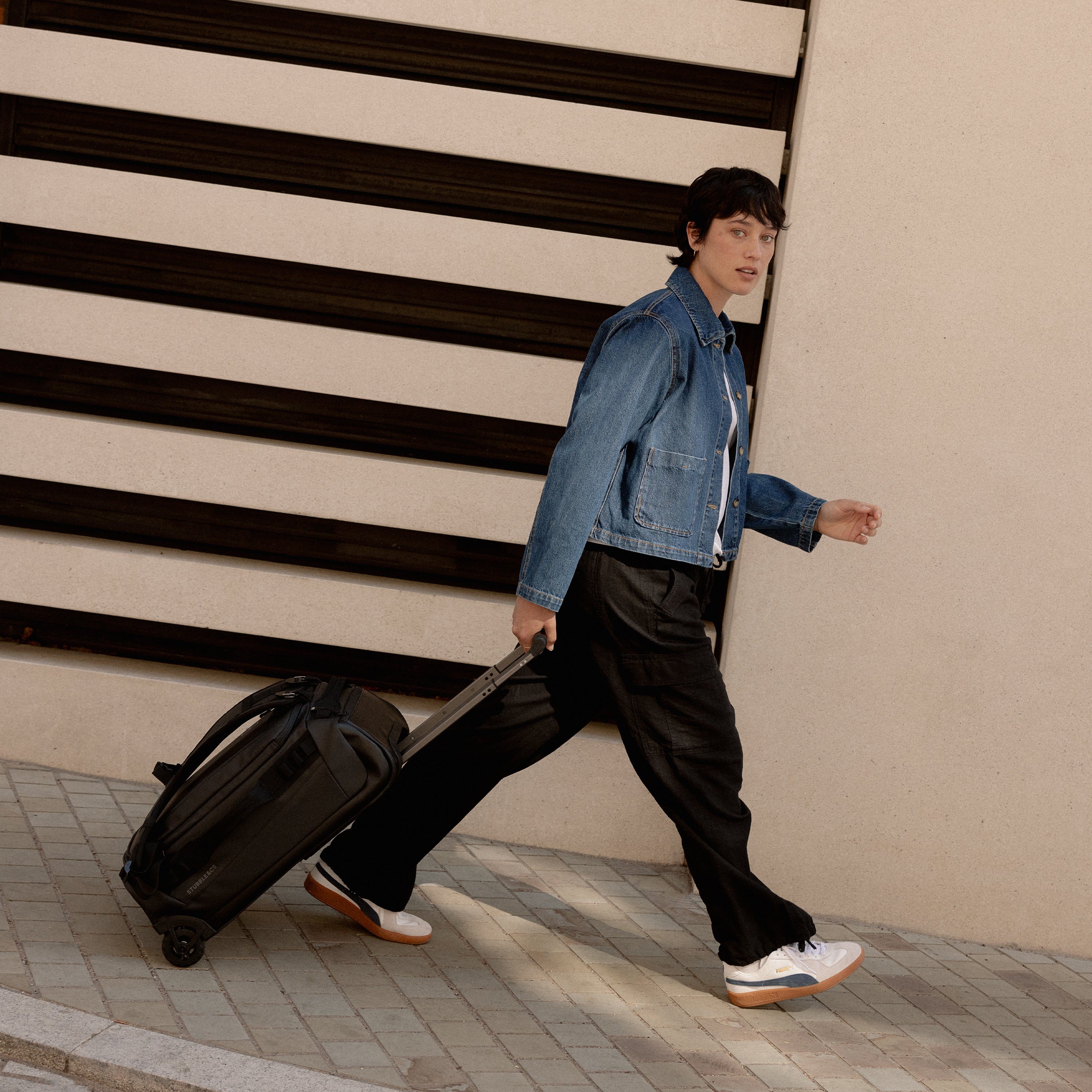 A woman walking down the street wheeling a Wheeled Kit bag 40L in All Black behind her